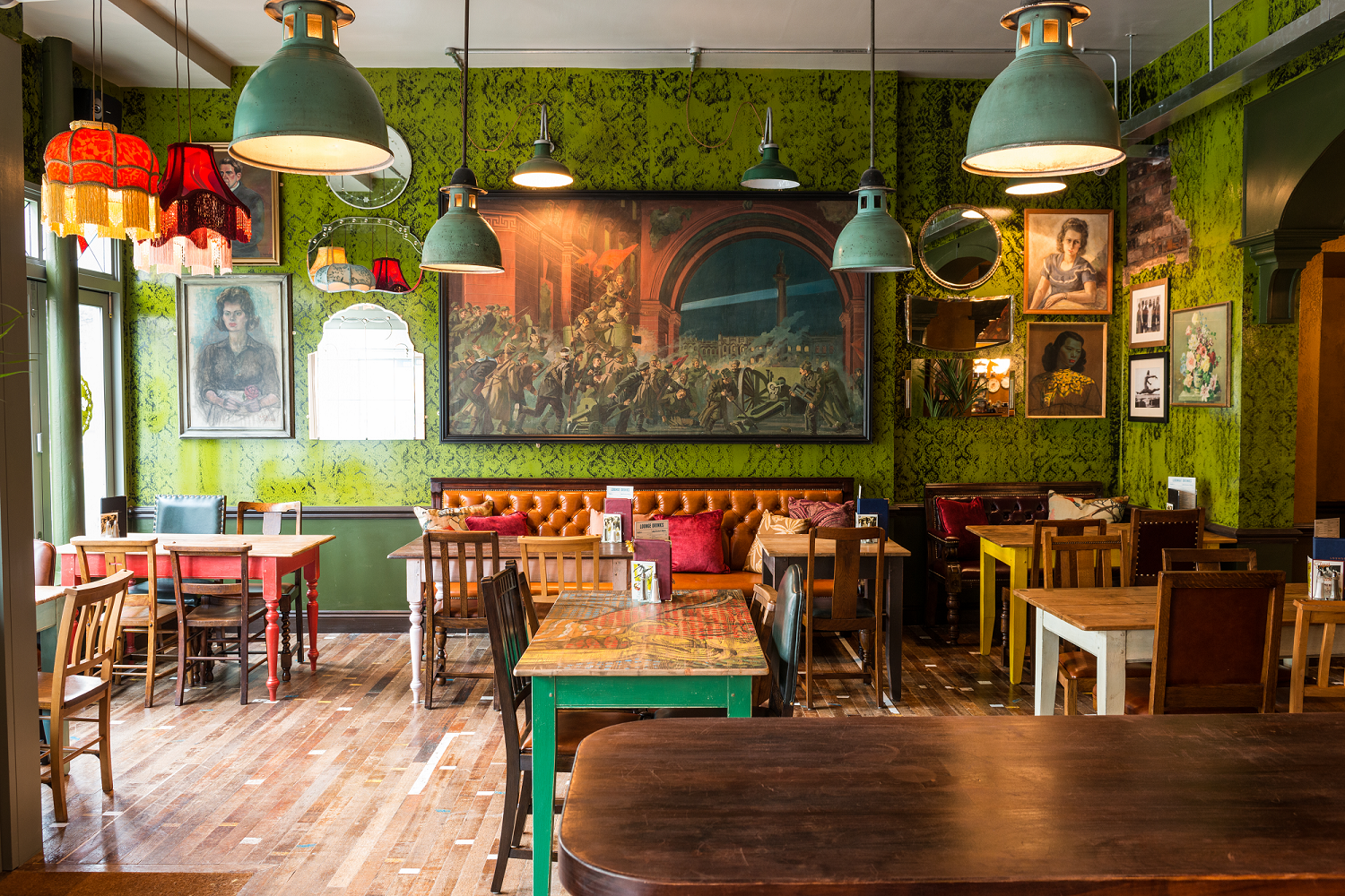 Colorful, eclectic café interior with green patterned walls, mismatched tables and chairs, vintage lamps, framed artwork, a playful Milo mural, and wooden floor, creating a cozy and artistic atmosphere.