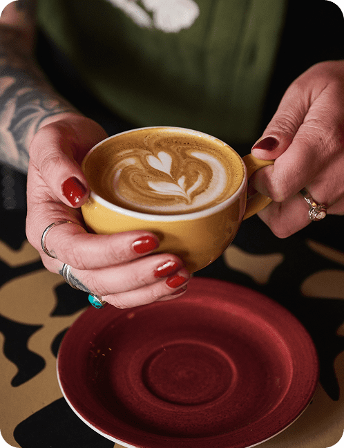 A person with red nail polish and rings holds a yellow cup of latte with heart-shaped latte art above a red saucer on a patterned table in Torino.